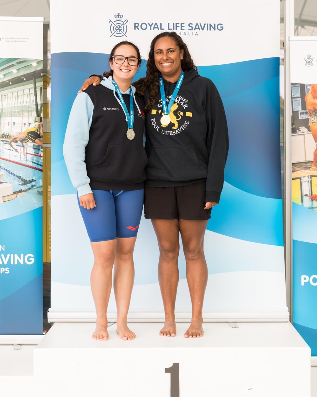 Kirra Dale and Leah Jones standing on the podium in shared 1st Place. Both are wearing medals around their necks.