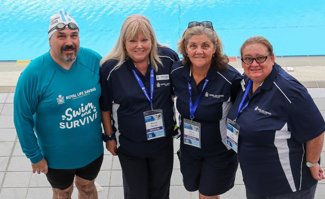 Four of the Royal Life Saving - Australia Pool Life Saving Officials, in their blue Royal Life Saving polo shirts, are standing in front of a swimming pool