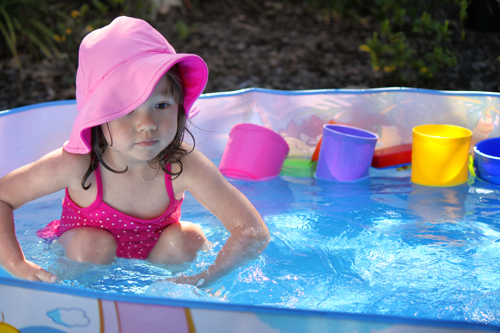 A young toddler sits in a bright blue portable pool. She wears a pink sun hat and behind her floats colourful pool toys.