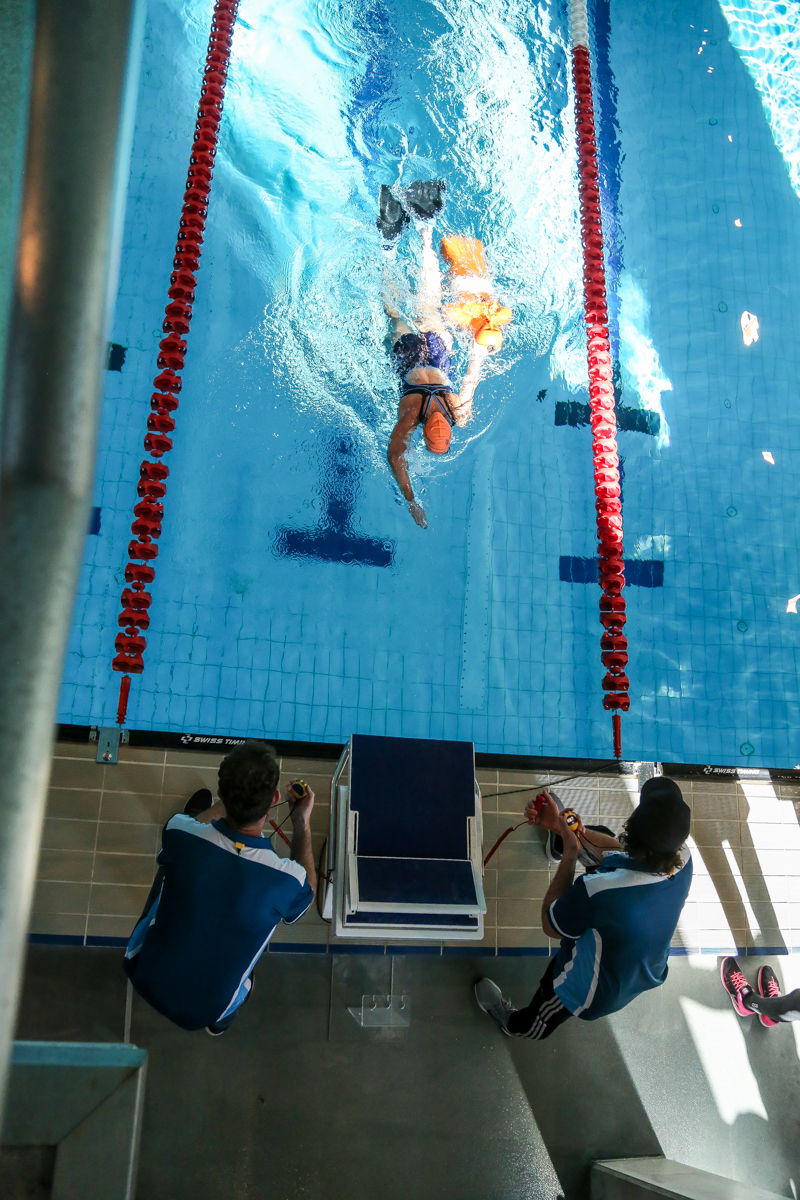 Two officials using timers to record the race time. A woman swimming carrying an orange manikin.