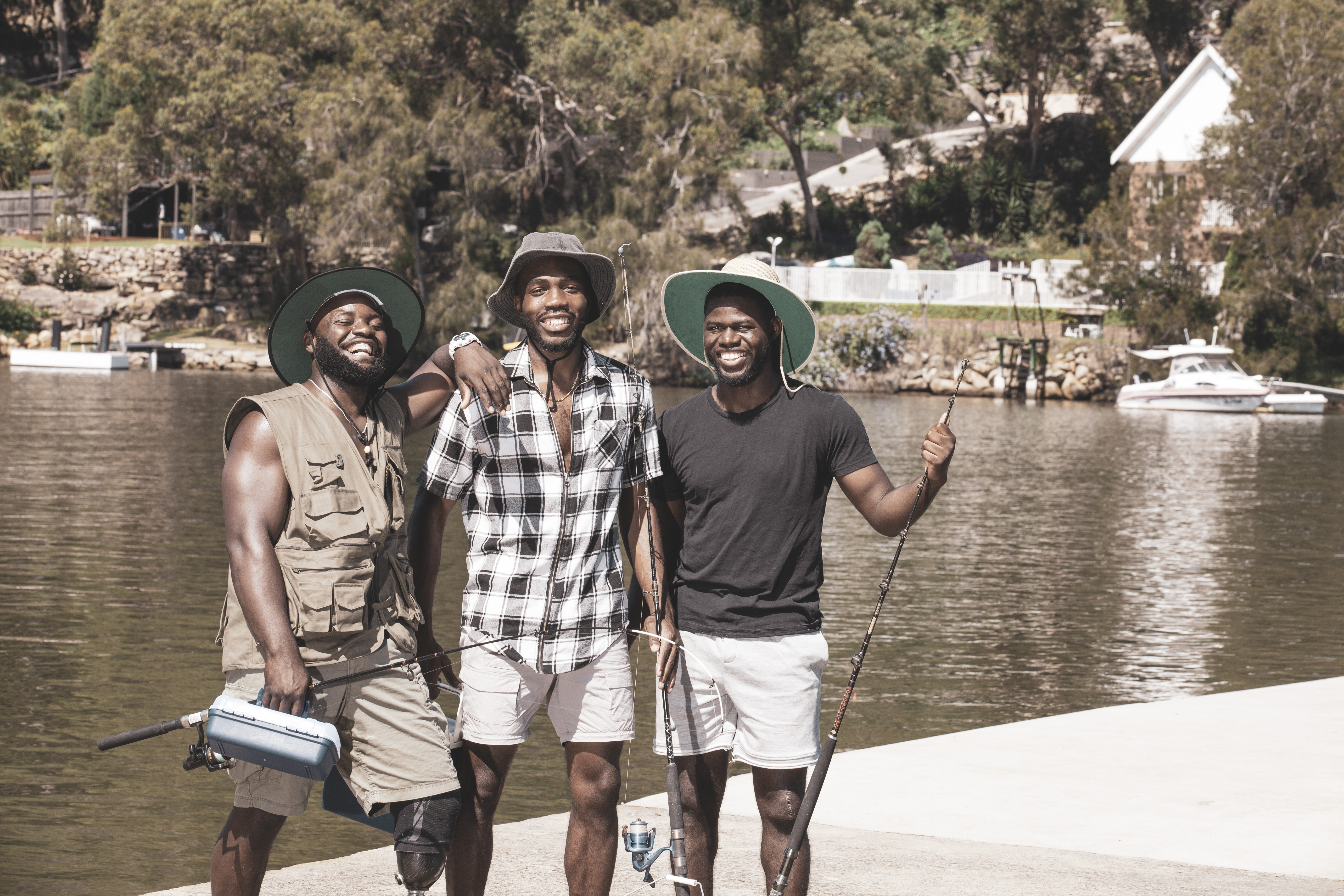 Three men stand in front of the water, wearing sun hats and holding fishing gear. They are smiling and leaning on each other's shoulders.